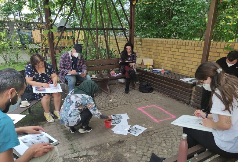 Menschen sitzen im Kreis, malen und zeichnen. Eine Frau hat ein Kopftuch. 