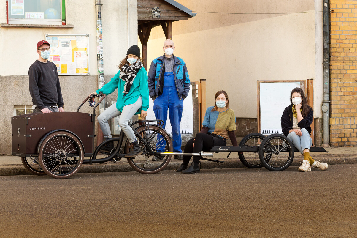 4 Menschen umringen ein Lastenrad und einen Fahrrad-Schwerlast-Anhänger. Alle tragen eine Corona-Maske.